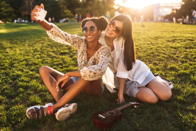 Красивые улыбающиеся девушки, сидящие на траве, счастливо фотографируют на мобильный телефон, проводя время вместе в городском парке