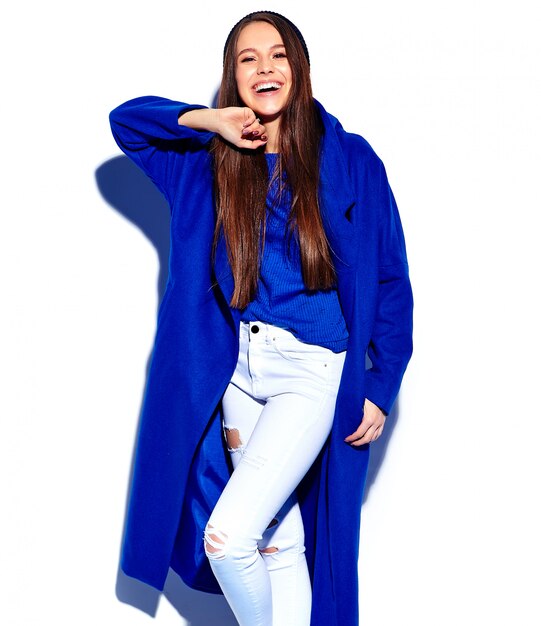 白で隔離されるスタイリッシュな青いオーバーコートで美しい笑顔ヒップスターブルネットの女性モデル