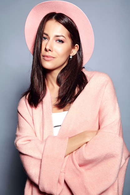 Beautiful smiling hipster brunette woman model in stylish pink overcoat and colorful hat posing on gray