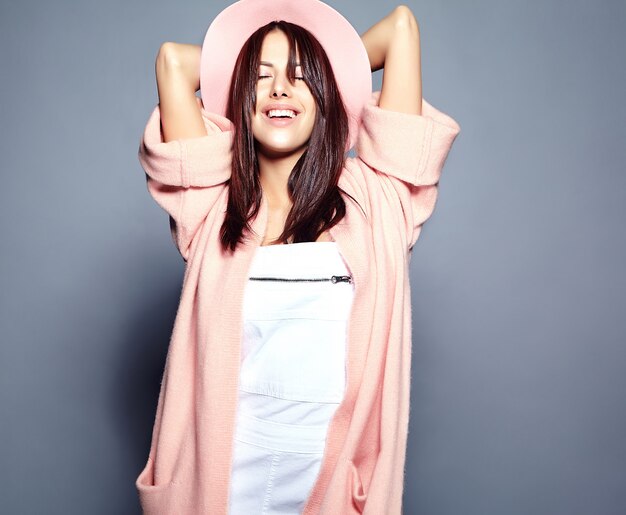 Beautiful smiling hipster brunette woman model in stylish pink overcoat and colorful hat posing on gray