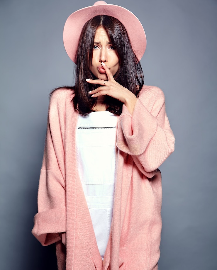 Free Photo beautiful smiling hipster brunette woman model in stylish pink overcoat and colorful hat posing on gray