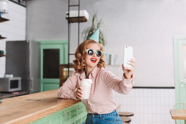 Beautiful smiling lady in sunglasses and birthday cap sitting at the bar counter with milkshake and taking cute photos on her cellphone in cafe