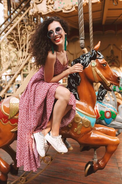 Beautiful smiling lady with dark curly hair in sunglasses and dress sitting on carousel horse while happily spending time in amusement park