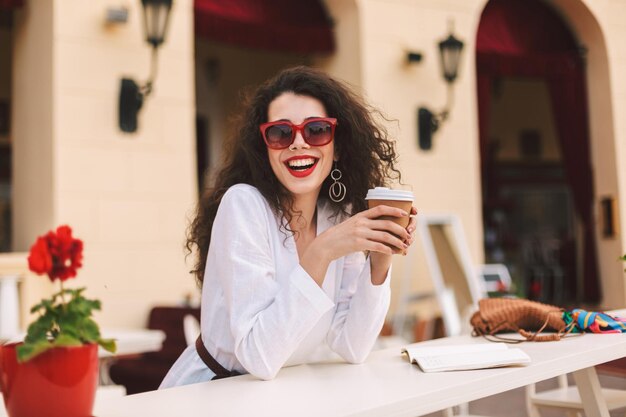 サングラスをかけた濃い巻き毛の美しい笑顔の女性がコーヒーを片手に立って、カフェの夏のテラスで時間を過ごしながらカメラを見て幸せに見ています