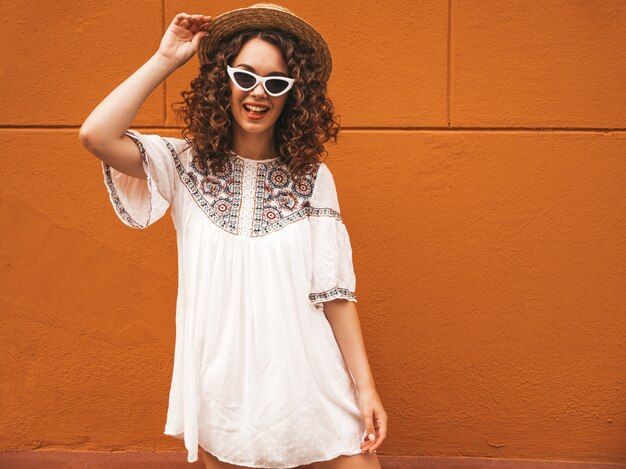 Красивая улыбающаяся модель с прической в афро-локонах, одетая в летнее белое платье и солнцезащитные очки