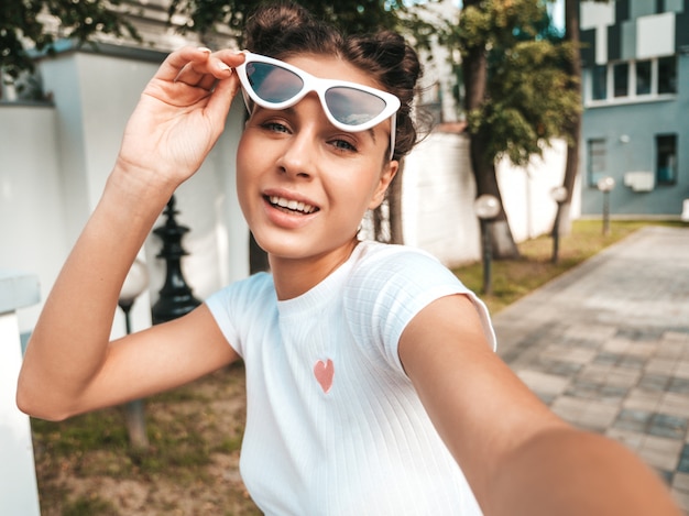 Beautiful smiling model with horns  hairstyle dressed in summer casual clothes.Sexy carefree girl posing in the street in sunglasses.Taking selfie self portrait photos on smartphone