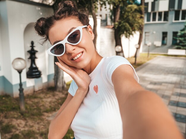 Beautiful smiling model with horns  hairstyle dressed in summer casual clothes.Sexy carefree girl posing in the street in sunglasses.Taking selfie self portrait photos on smartphone