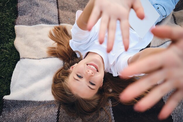 Beautiful smiling teenage girl lying on plaid while happily pulling hands up to camera on picnic in park