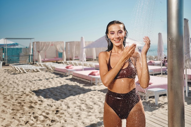 Beautiful smiling wet brunette girl in swimsuit taking beach shower happily looking in camera on seaside