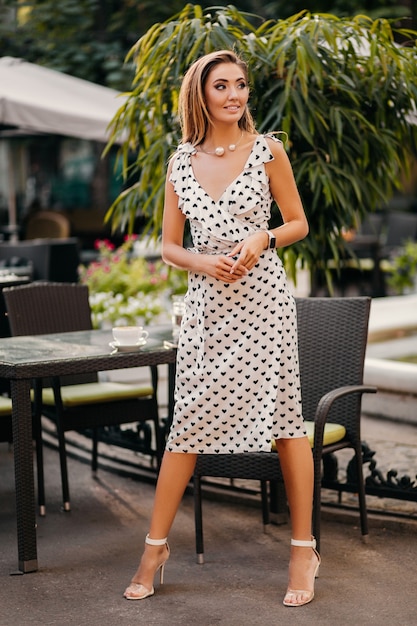 Beautiful smiling woman dressed in white printed dress in romantic style posing in street cafe on sunny day