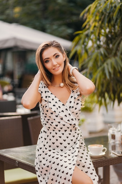 Красивая улыбающаяся женщина, одетая в белое платье с принтом в романтическом стиле, позирует в уличном кафе в солнечный день