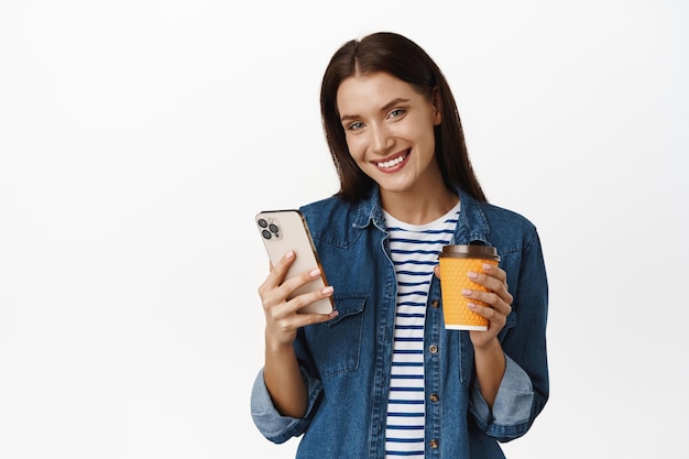 Beautiful smiling woman drinking coffee from yellow takeaway cup of coffee shop, holding smartphone, using mobile phone and looking happy at camera, white background