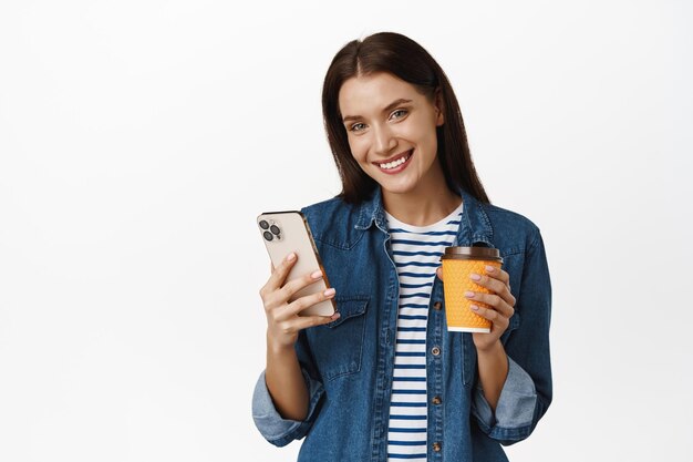 Красивая улыбающаяся женщина пьет кофе из желтой чашки на вынос в кофейне, держит смартфон, пользуется мобильным телефоном и счастливо смотрит в камеру, белый фон