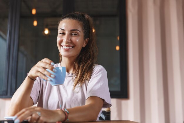 집 테라스에서 차를 마시고 컵을 들고 티셔츠 차림으로 커피 테이블에 앉아 카메라를 바라보며 행복한 미소를 짓고 있는 아름다운 여성