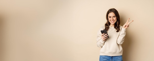 Beautiful smiling woman showing peace sign holding mobile phone standing over beige background