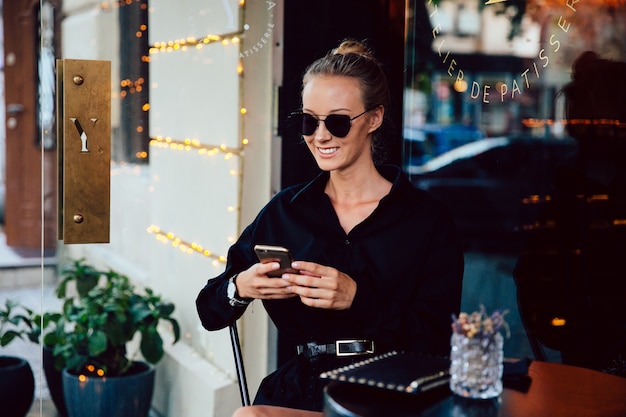 Beautiful smiling woman in sunglasses and black elegant coat, using a mobile phone