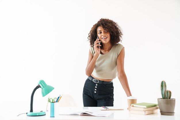 Beautiful smiling young african businesswoman wearing casual clothing standing at the desk isolated over white wall, talking on mobile phone
