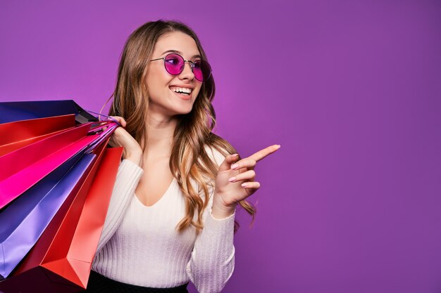 Красивая улыбающаяся молодая белокурая женщина, указывающая в солнцезащитных очках, держащая хозяйственные сумки и кредитную карту на розовой стене