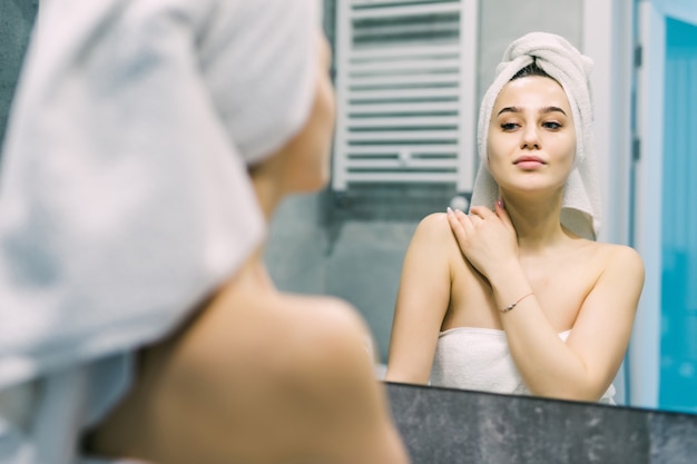 Beautiful smiling young woman in bathrobe and towel on head looking at mirror in bathroom