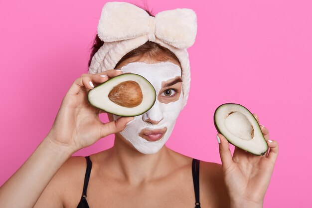 Beautiful spa woman with facial mask on face and holding halves of avocado in hands, looking at camera, doing cosmetology procedures at home, wearing hair band with bow.