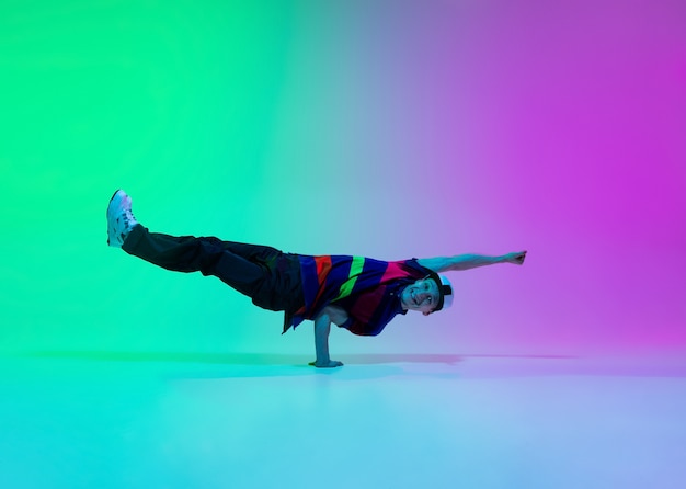 Beautiful sportive boy dancing hip-hop in stylish clothes on colorful gradient wall at dance hall in neon light. Youth culture, movement, style and fashion, action. Fashionable bright portrait.