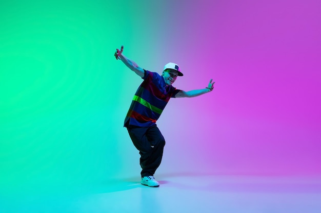 Free photo beautiful sportive boy dancing hiphop in stylish clothes on colorful gradient background at dance hall in neon light