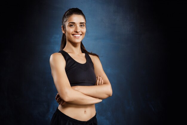 Beautiful sportive girl posing with crossed arms over dark wall.