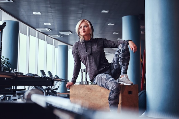 Beautiful sportive girl with blonde hair wearing hoodie sitting on a wooden box and looking sideways in the gym