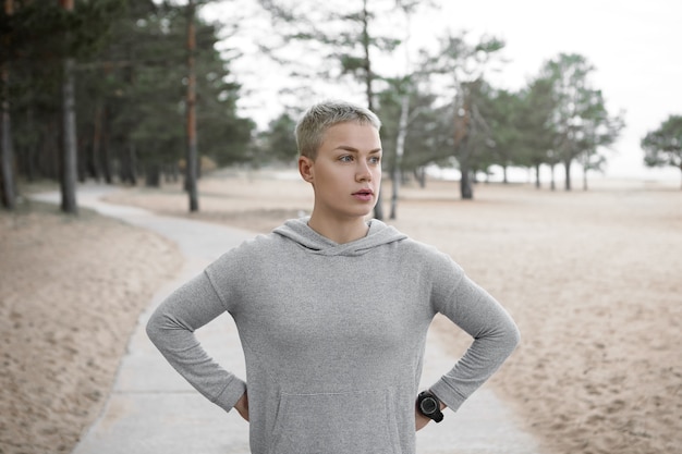 Beautiful sporty young short haired blonde woman having tired look, catching breath after morning run, holding hands on her waist, posing on paved trail against sandy beach and pine trees background