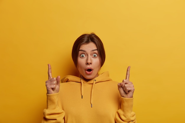 Beautiful stupefied European woman indicates upwards, introduces new product, gasps from wonder, has bugged eyes, invites to click promo banner, wears hoodie, isolated over yellow wall