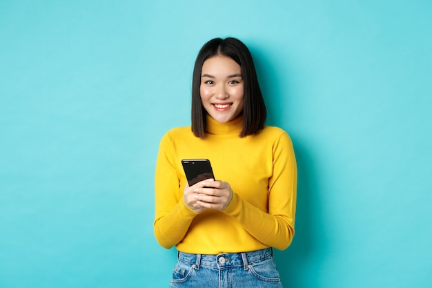파란색 배경 위에 서서 휴대전화로 온라인 쇼핑을 하는 아름답고 세련된 아시아 여성