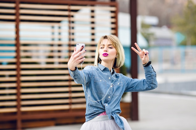 Beautiful stylish blond girl is having fun and makes selfie with a kiss face and with victory sign fingers with striped brown wooden balks behind