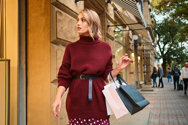 Beautiful stylish blond girl in knitted sweater with shopping bags dreamily walking on city street