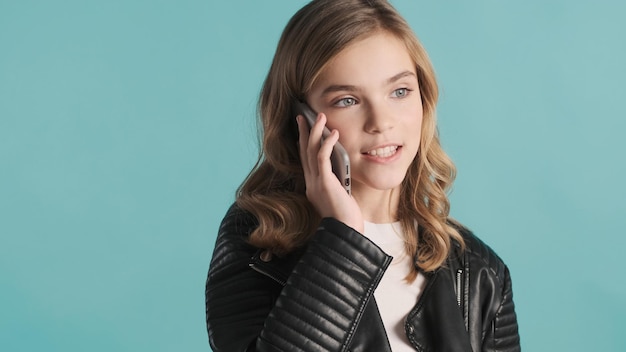 Beautiful stylish blond teenage girl dressed in leather jacket talking on phone over blue background. Distant communication