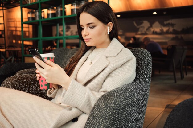 モダンなカフェで休んでいる携帯電話を自信を持って使用しているワイヤレスイヤホンの美しいスタイリッシュな実業家