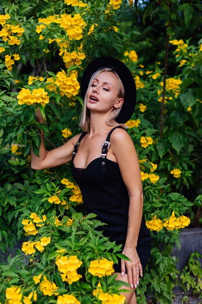 노란색 타이어 꽃으로 둘러싸인 공원에서 검은 드레스와 클래식 모자에 아름 다운 세련 된 백인 행복 한 여자