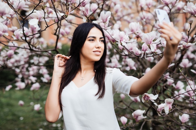 Beautiful stylish caucasian woman making selfie in blossom magnolia garden. Bottom view