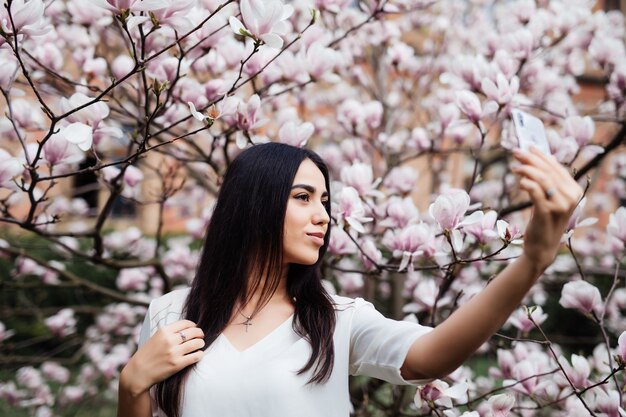 花のマグノリアの庭で自分撮りを作る美しいスタイリッシュな白人女性。春の時間