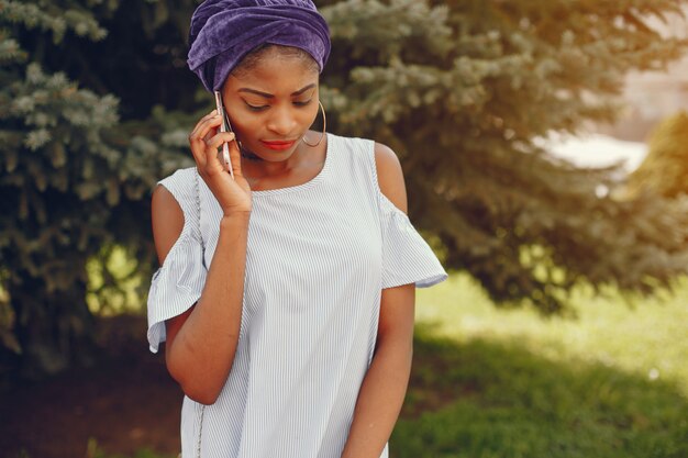 A beautiful and stylish dark-skinned girl walks in a sunny summer park with phone