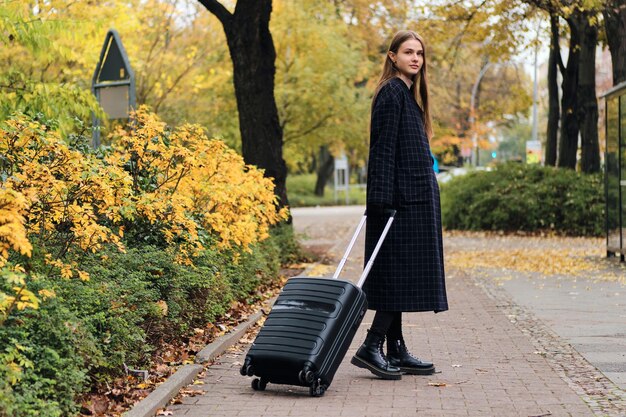 Beautiful stylish girl in coat with luggage bag intently looking in camera in city park