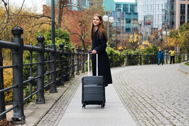 Beautiful stylish girl in coat with luggage bag joyfully looking in camera on city street