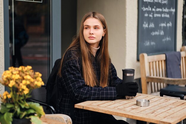 居心地の良いストリートカフェで思慮深く目をそらしているサーモカップを保持している美しいスタイリッシュな女の子