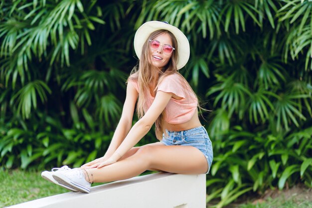 Beautiful stylish girl sitting on white fence in tropical park with stretched legs.