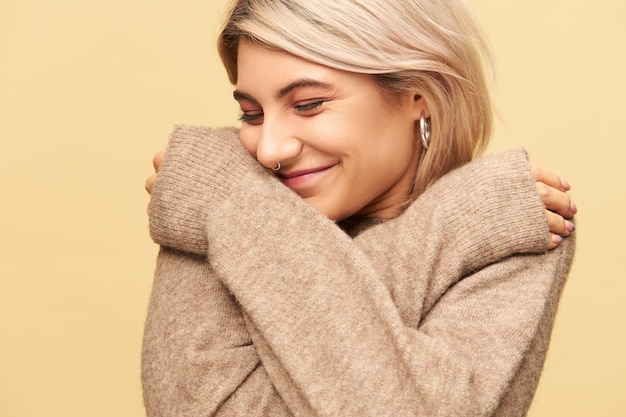 Beautiful stylish girl with facial piercing being in good mood posing isolated with arms crossed on her chest, wearing cozy cashmere sweater with long sleeves, warming up on cold December day