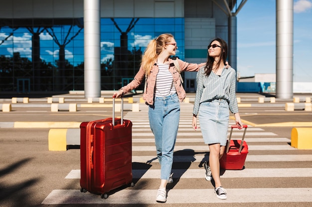 Красивые стильные девушки в солнечных очках счастливо гуляют по пешеходной полосе с красными чемоданами и аэропортом на заднем плане