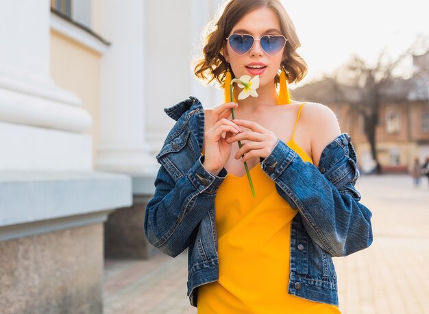 Красивая стильная хипстерская женщина позирует, уличная мода, цветок в руках, желтое платье, джинсовая куртка, стиль бохо, весенне-летний модный тренд, улыбка, модные синие солнцезащитные очки, улыбка, солнечно, аксессуары