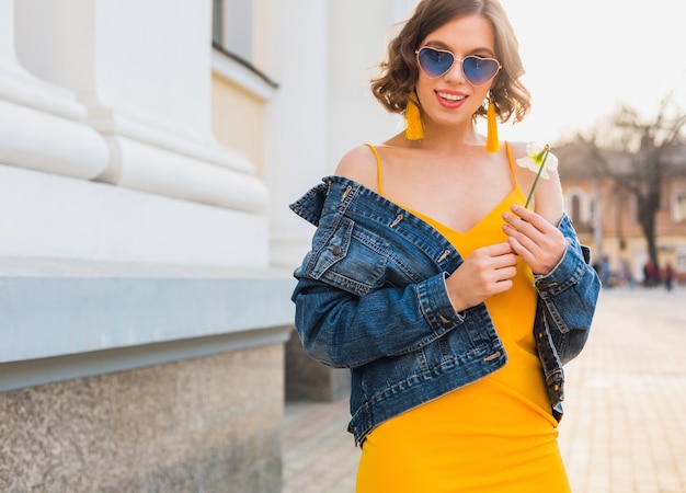 Красивая стильная хипстерская женщина позирует, уличная мода, цветок в руках, желтое платье, джинсовая куртка, стиль бохо, весенне-летний модный тренд, улыбка, модные синие солнцезащитные очки, улыбается, солнечно