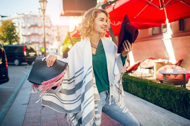 Beautiful stylish lady walking in street in cape holding handbag, fashion accessories, spring street style trend, smiling