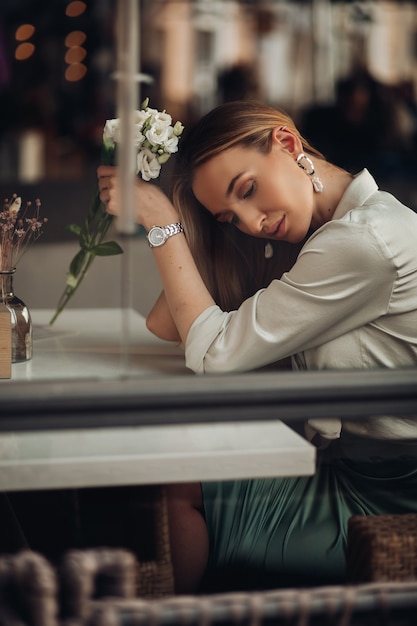 Beautiful stylish romantic young girl wearing evening clothes and jewelry sitting, dreaming with flower in hand in cafe. High quality photo