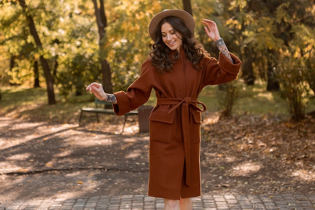 무료 사진 따뜻한 갈색 코트, 가을 유행 패션 거리 스타일을 입고 공원에서 산책하는 곱슬 머리를 가진 아름 다운 세련 된 웃는 마른 여자